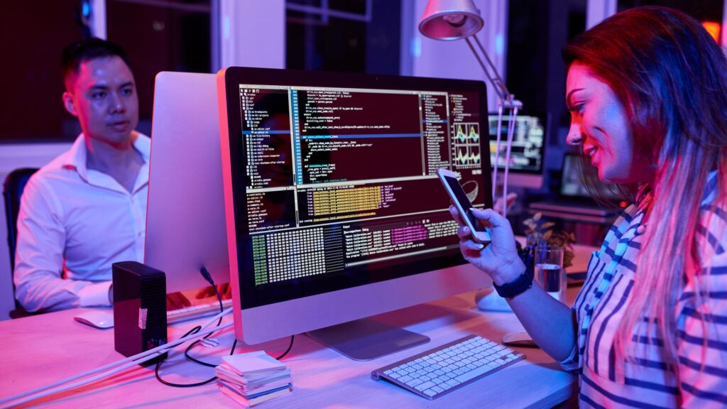 Lächelnde Softwareentwicklerin, die am Schreibtisch sitzt und mit Freunden SMS schreibt, anstatt zu programmieren